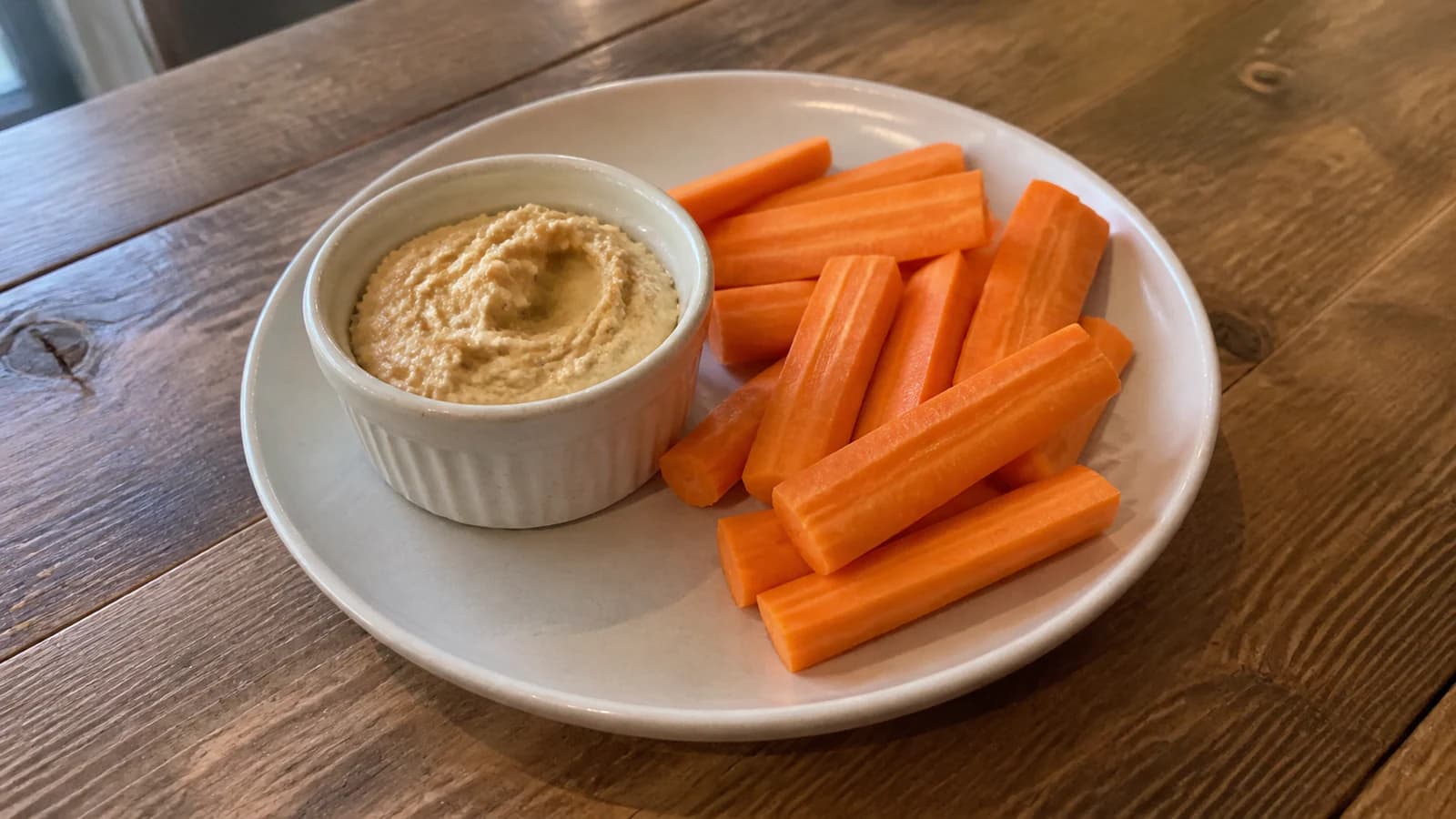 Fresh carrot sticks beside a small ceramic ramekin of plain hummus